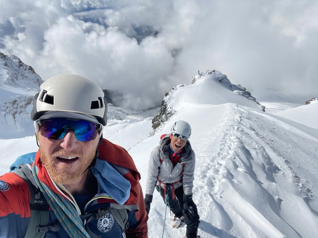Zwei Bergsteiger im Schnee, einer davon lächelt in die Kamera, während die andere Person hinter ihm auf einem schmalen Grat steht. Im Hintergrund sind Wolken und eine Berghütte sichtbar.